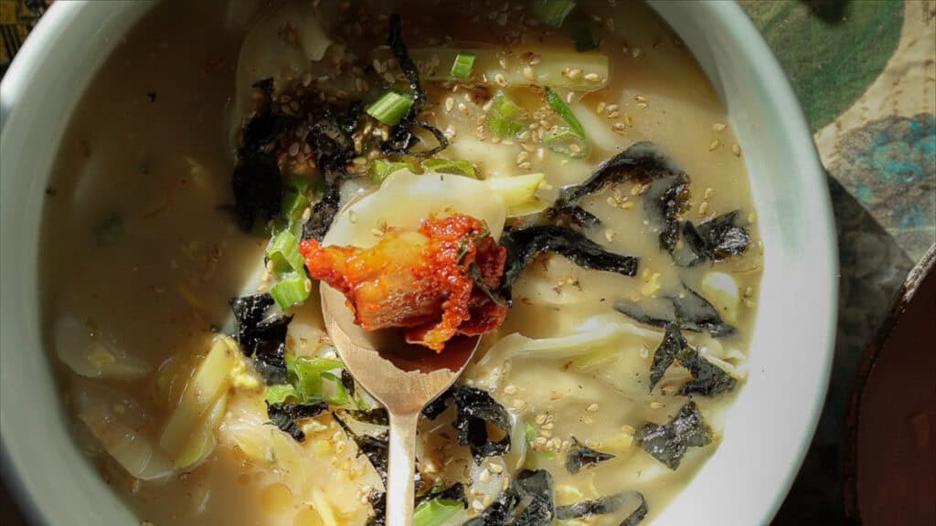 A bowl of savory vegan rice cake soup with sliced seaweed, chopped green onions, sesame seeds, and a wooden spoon holding a dollop of spicy sauce. The soup has a creamy broth with thin noodles, creating a hearty and flavorful dish.